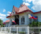 Looking at al old wooden house painted white with red roof and white iron fencing and a red, white, yellow and blue flags in front of it.