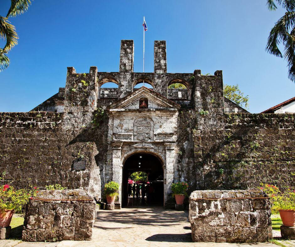 Fort San Pedro a historic stone fortress with arch entrance, flag on top, set against a clear blue sky. Greenery and potted plants surround the entrance.