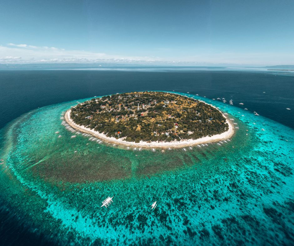 Aerial view of a lush круг? no english. Need English. Aerial view of a lush circular tropical island ringed by turquoise reef, with small boats on calm blue sea.