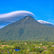Green mountain with a unique cloud cap, blue sky backdrop, lush vegetation, and a colorful village at the base. Serene atmosphere.