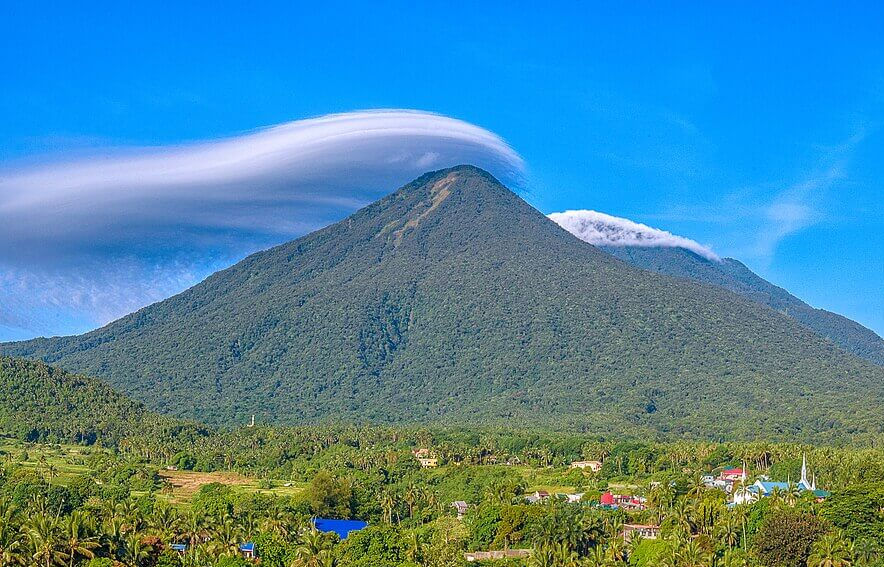Green mountain with a unique cloud cap, blue sky backdrop, lush vegetation, and a colorful village at the base. Serene atmosphere.