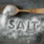 Wooden spoon filled with coarse salt on a gray surface. Salt is spilled, forming the word "SALT" in bold, white letters.