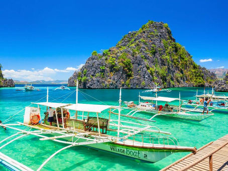 Boats docked in turquoise water, vibrant greenery on rocky island under clear blue sky. "Village Boat" text visible. Calm, sunny scene.