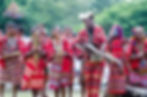 People in vibrant red traditional attire, playing instruments and smiling in a lush outdoor setting, convey joy and cultural celebration.