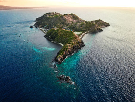 Aerial view of a lush tropical island in blue sea at sunset, with small boats offshore and calm waters.