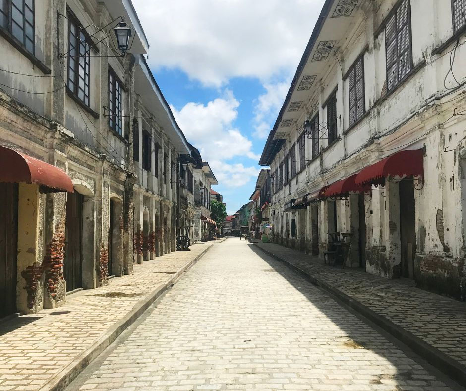 Cobblestone street between historic, weathered buildings with red awnings. Bright blue sky and scattered clouds in the background. Peaceful vibe.