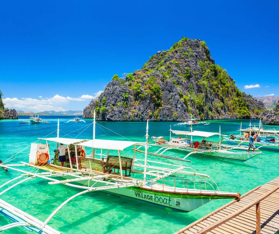 Boats docked on turquoise water with a lush green island backdrop under a clear blue sky. A wooden pier is in the foreground.