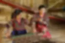 Looking at two women with their traditional red, black, yellow and white tribal dress weaving fabric.