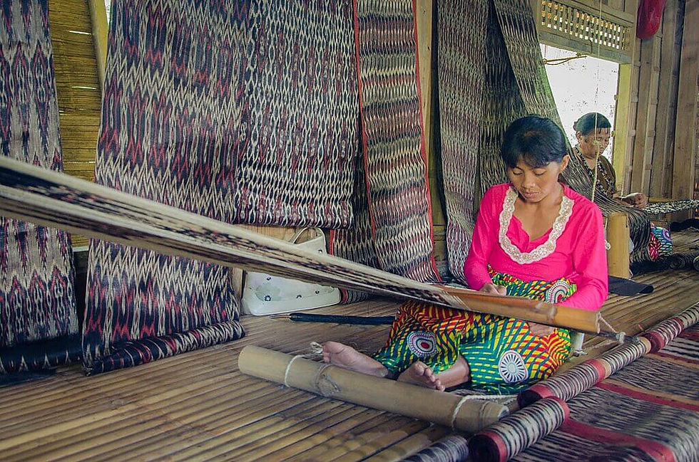 Two T'boli woman Dreamweaver's, weave using wooden looms in a bamboo room. Brightly patterned fabric surrounds them. One wears a pink blouse, focused and serene.