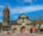 Looking at a large old gray stone church building with red paving stone leading to the church on a clear blue-sky.