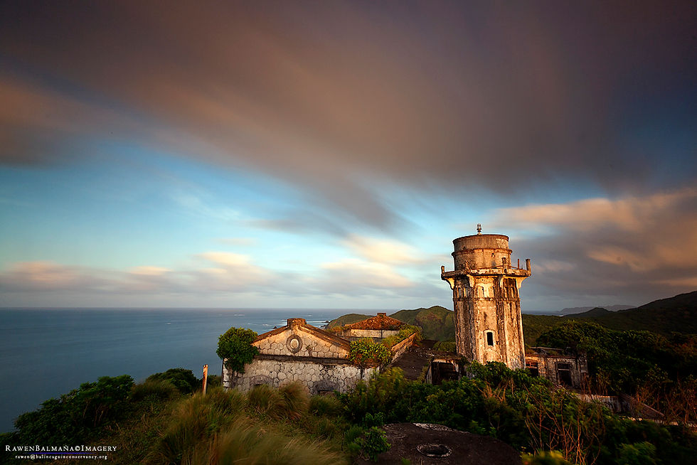 Old stone lighthouse and building on a lush hill with ocean view. Dramatic sky in warm tones, creating a serene, timeless mood.