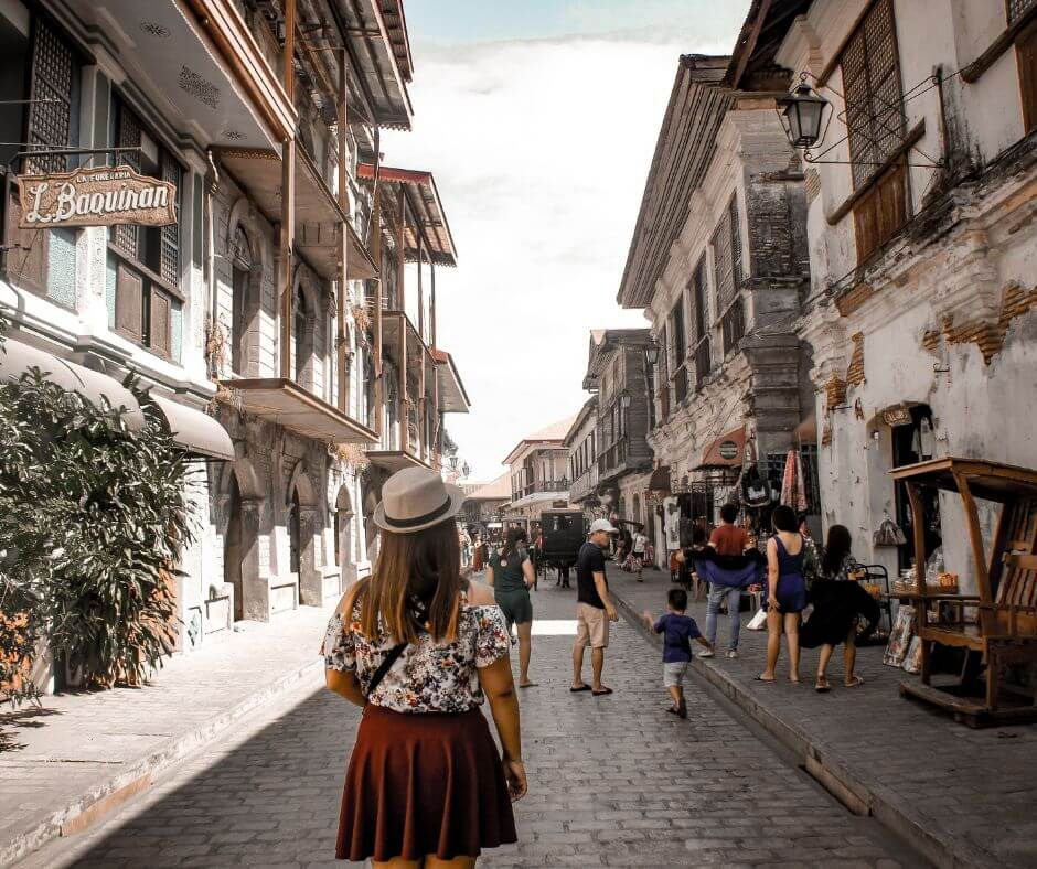 A woman in a hat walks down a cobblestone street lined with historic buildings. People are strolling, and a sign reads L. Bauinan.