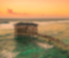Wooden stilt hut on ocean, crowded with people at sunset. Orange sky, waves crashing, visitors walking on a wooden path over water.