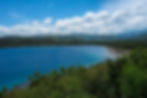 Blue lake with a small boat, lush green hills, and a partly cloudy sky. Dense greenery lines the foreground, evoking a serene mood.