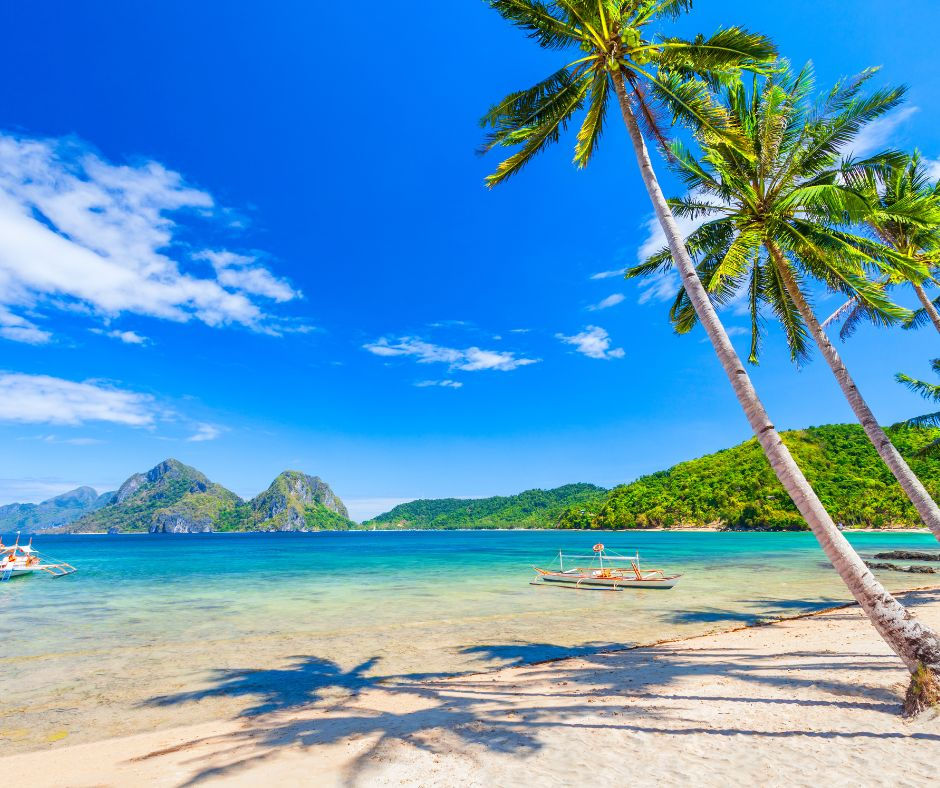 Tropical beach with leaning palm trees, clear turquoise water, small boats, green islands, and a bright blue sky.