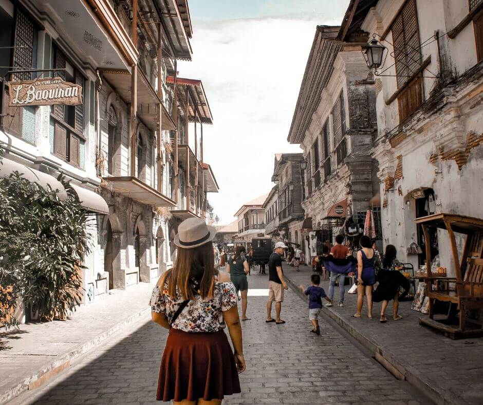 A woman in a hat walks down a cobblestone street with historical buildings in Vigan Ilocos Sur. People and shops line the romantic, sunlit path. Mood is lively.