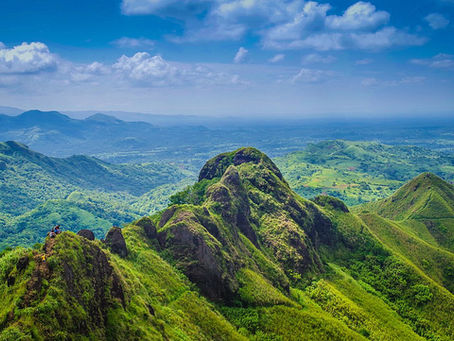 Mount Batulao in Batangas a lush green mountain under a vibrant blue sky with scattered clouds. Two people sit on a peak, enjoying the expansive view.
