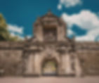 Historic stone gate with intricate carvings under a bright blue sky with fluffy clouds. Lush greenery is visible beyond the archway.