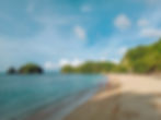 Sandy beach with two people walking along the shore. Green islands in the background, blue sky with clouds, and lush greenery on the right.