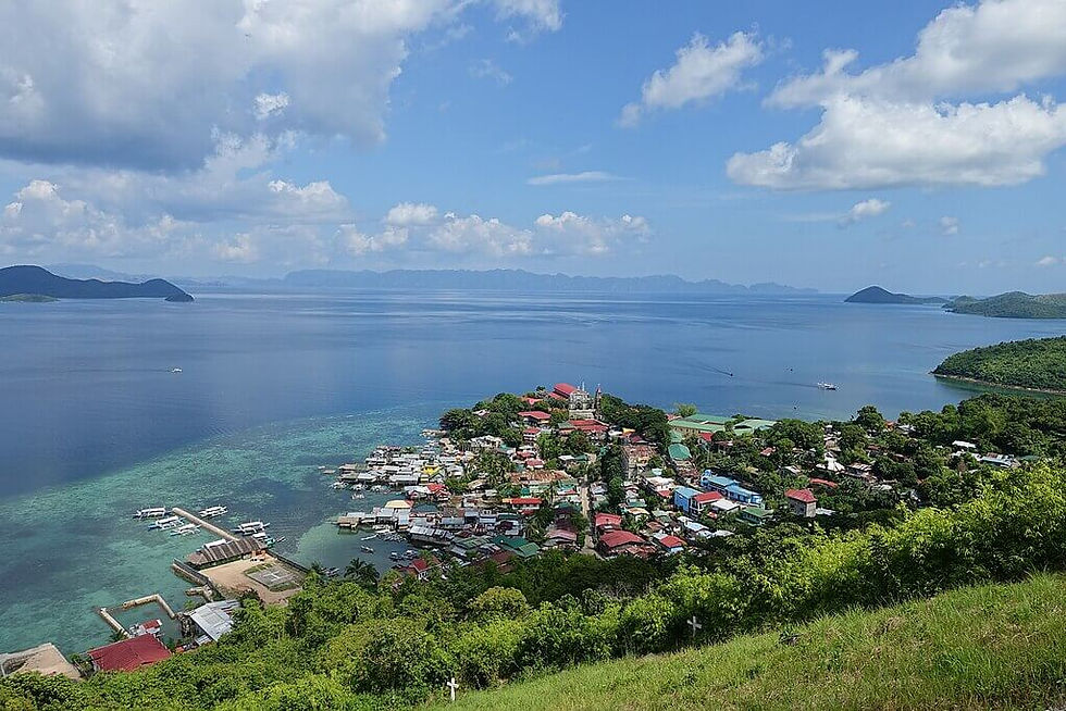 A coastal town with colorful rooftops surrounded by lush greenery, overlooking a calm sea under a bright blue sky with clouds.