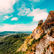 Rugged mountains with lush greenery and rocky cliffs under a bright blue sky with scattered clouds; expansive landscape view.