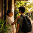 Smiling woman touches a young man’s shoulder outside a rural home, with a backpack on his back and warm tropical greenery behind them.