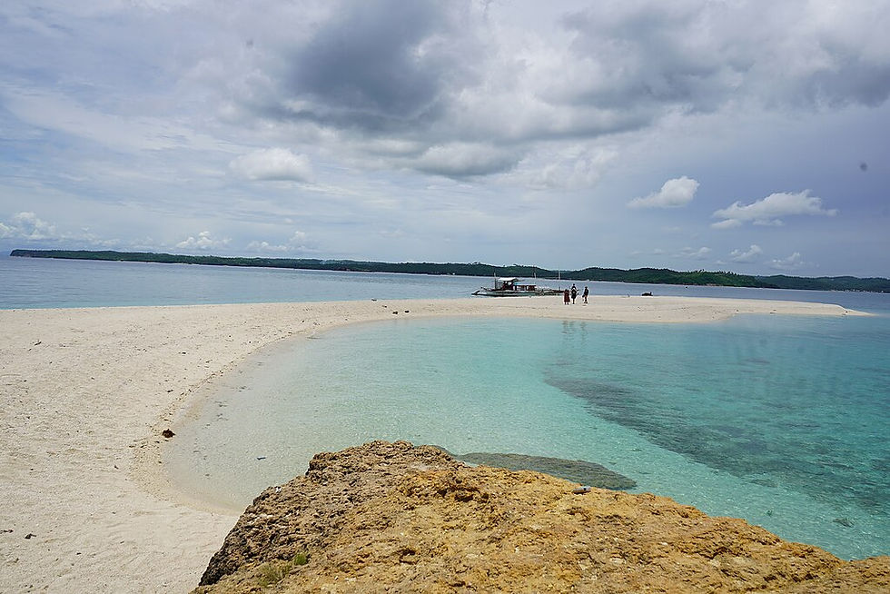 Sandy beach with clear turquoise water, small group near a boat, cloudy sky, and distant green hills. Peaceful and serene setting.