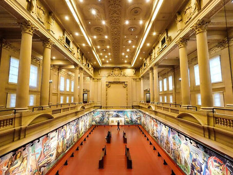Ornate museum hall with red floor and colorful murals, a few people walking beneath tall columns and bright ceiling lights.