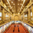 Ornate museum hall with red floor and colorful murals, a few people walking beneath tall columns and bright ceiling lights.
