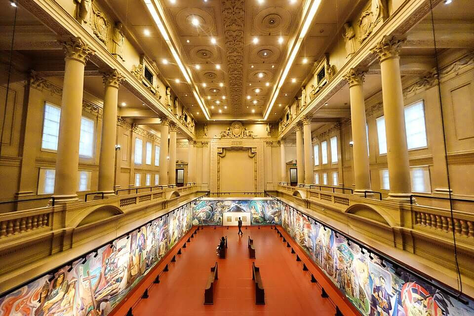 Ornate museum hall with red floor and colorful murals, a few people walking beneath tall columns and bright ceiling lights.