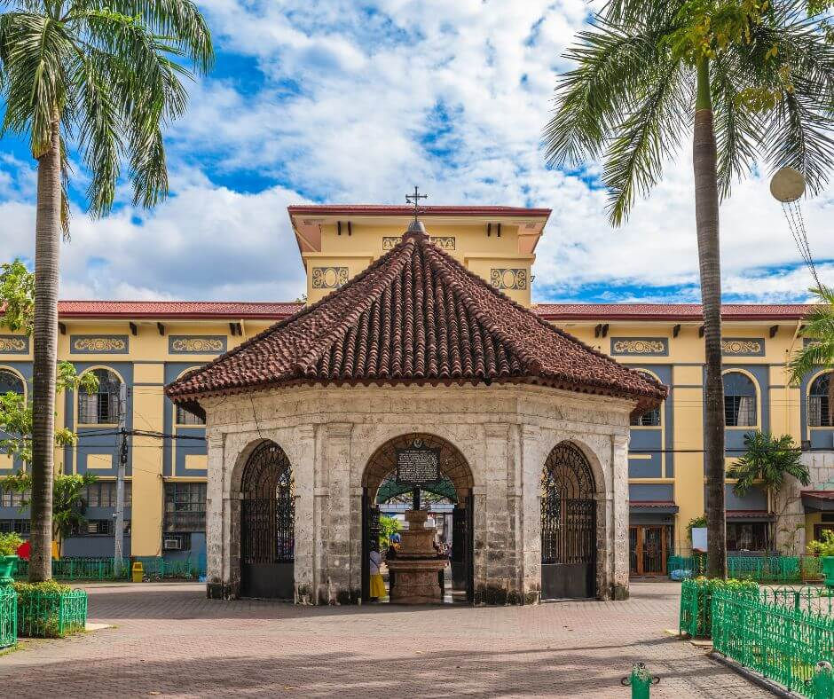 Magellan's Cross Pavilion in Cebu City a historic stone pavilion with red-tiled roof and arches, surrounded by palm trees. Clear sky and ornate building in the background.