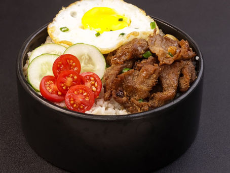 Black bowl of rice topped with fried egg, beef, cucumber slices, and cherry tomatoes on a dark background