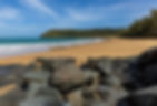 Looking at dark brown boulder rocks and golden sand beach with blue water lined with lush greenery under blue skies.