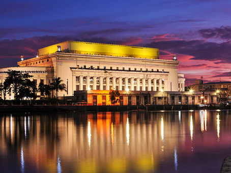 Manila Central Post Office a historic building with columns is illuminated at sunset. Its reflection shimmers in a calm river. Sky is vibrant with purple and orange hues.