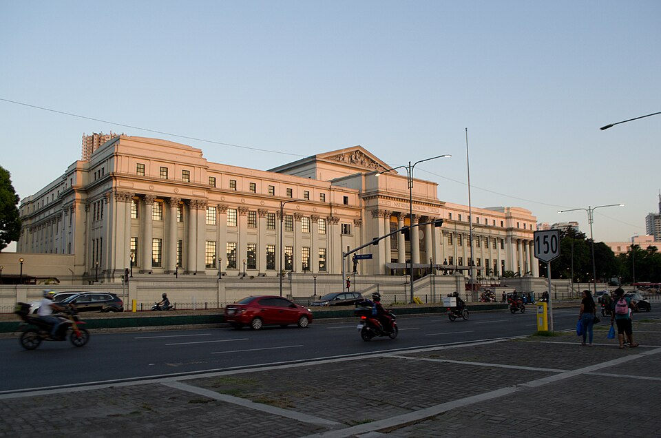 Neoclassical museum building at dusk beside a busy street with motorcycles, cars, pedestrians, and a 150 speed limit sign.
