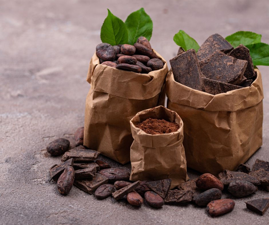 Brown paper bags with cocoa beans, chocolate chunks, and powder on a rough surface. Green leaves add a fresh touch.