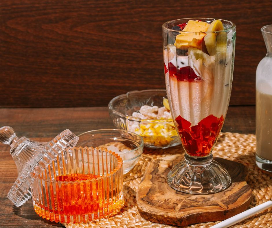 A colorful dessert in a tall glass with red jelly chunks, topped with fruit, on a rustic woven mat. Nearby, glass bowls are filled with condiments.
