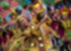 Dancer in vibrant, golden costume holds a statue, surrounded by colorful performers. Festive mood with intricate headdresses and patterns.