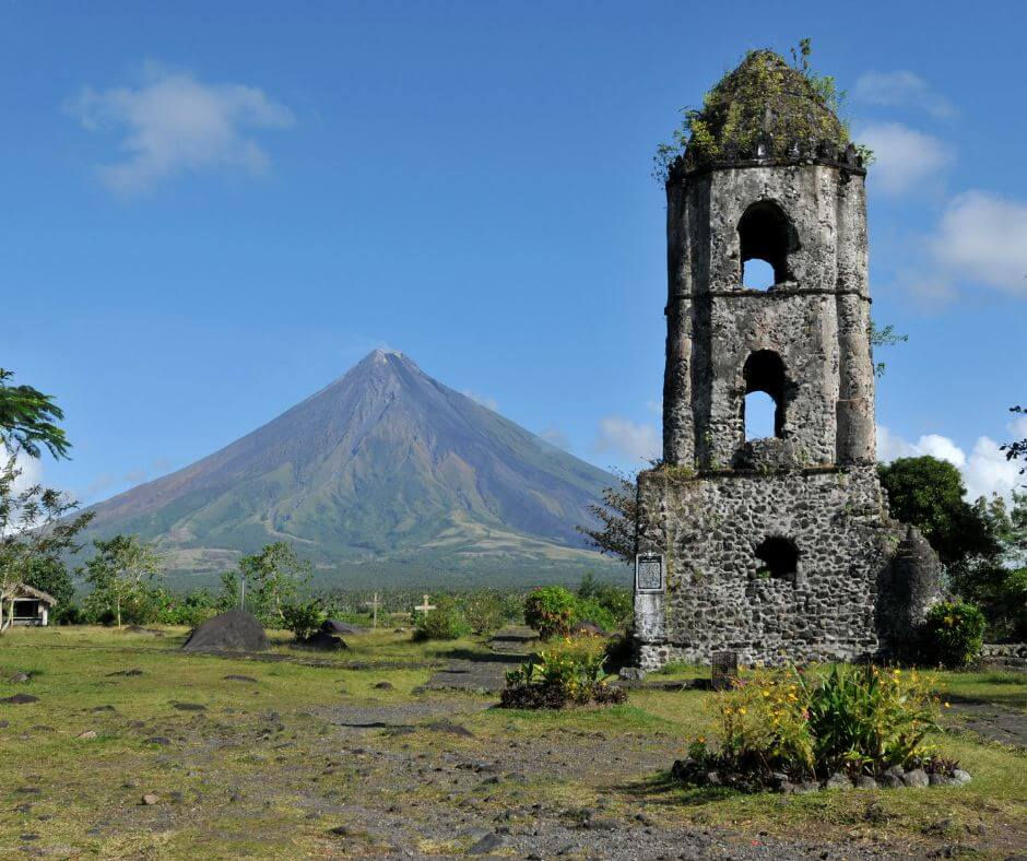 Mayon Volcano, a stone bell tower stands in lush greenery with Mayon Volcano in the background under a clear blue sky. Bright, vibrant landscape.