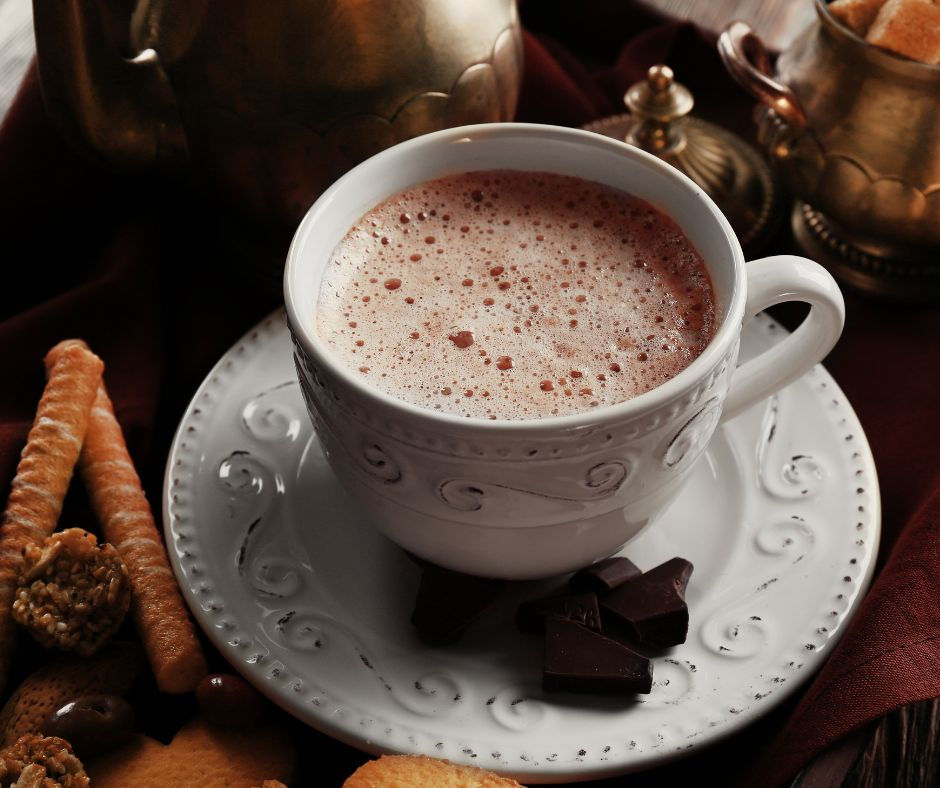 White cup of hot chocolate on a saucer, surrounded by assorted cookies and chocolate pieces. Vintage setting with a warm, cozy feel.