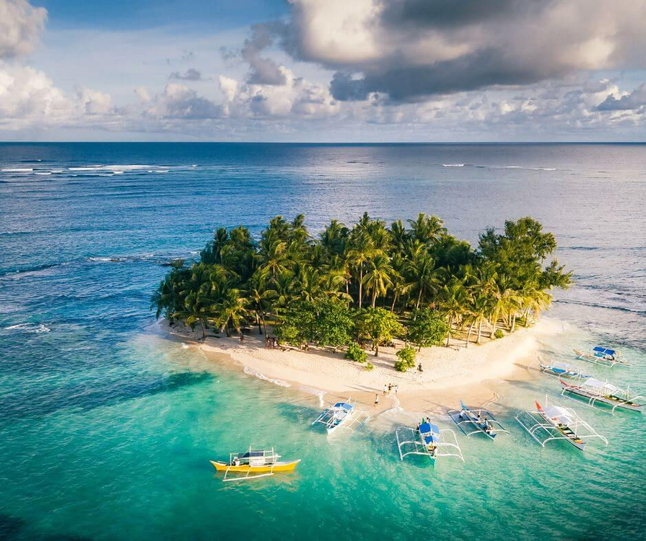 Tropical island with palm trees, turquoise water, outrigger boats, and a few people on a sandy beach under cloudy sky.