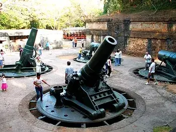 Battery Way Corregidor Island, people explore a historic site with large, green cannons on concrete platforms, surrounded by stone walls and greenery under a sunny sky.