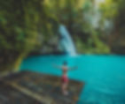 A man in red shorts stands on a bamboo raft with arms outstretched, facing a turquoise pool and waterfall in a lush forest setting.