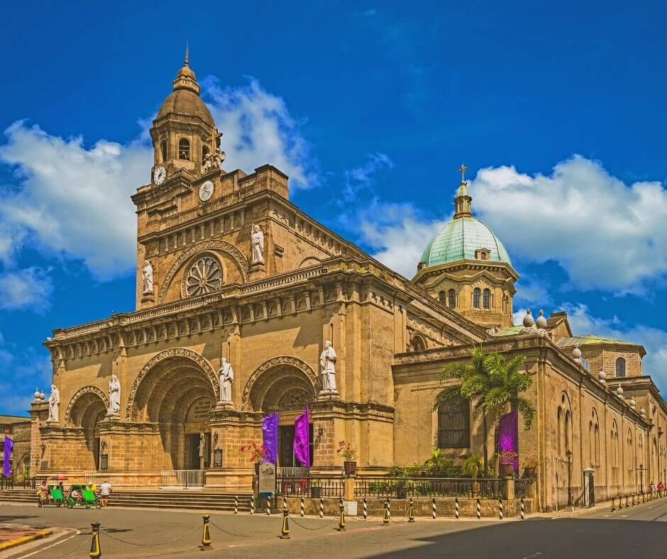 The Manila Cathedral a historic cathedral with ornate carvings and statues stands under a blue sky with fluffy clouds. Purple banners hang along the street.