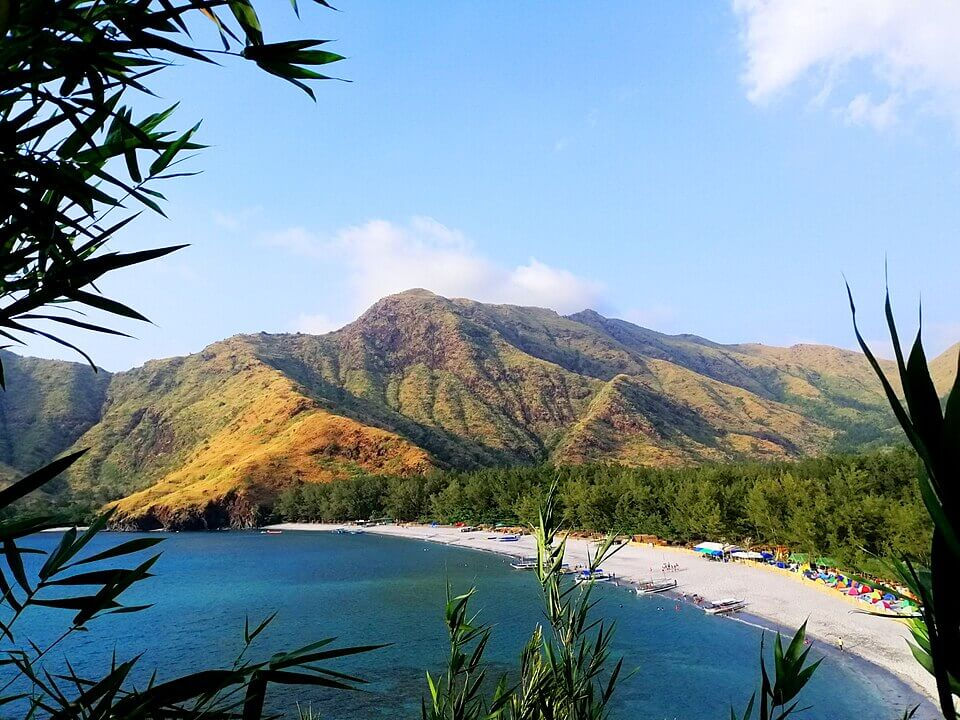 Tropical bay with blue water, sandy beach, green mountains, and bamboo leaves framing a bright, sunny sky.