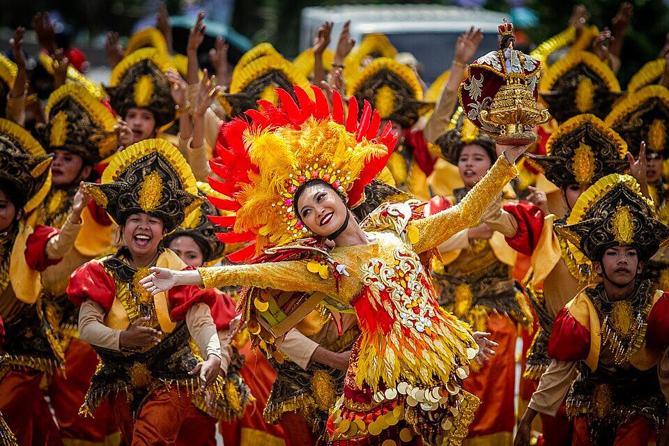 Dancers in vibrant yellow and red costumes perform joyfully in a lively street festival, adorned with ornate headdresses and bright feathers.