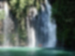 People in orange vests stand under a large waterfall surrounded by lush greenery. The water splashes into a clear, emerald pool below.