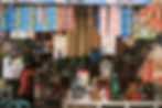 Sari-Sari Store, a small shop with hanging snacks and colorful detergent packets. A person in an orange shirt tends to items. Shelves are stocked with bottles.