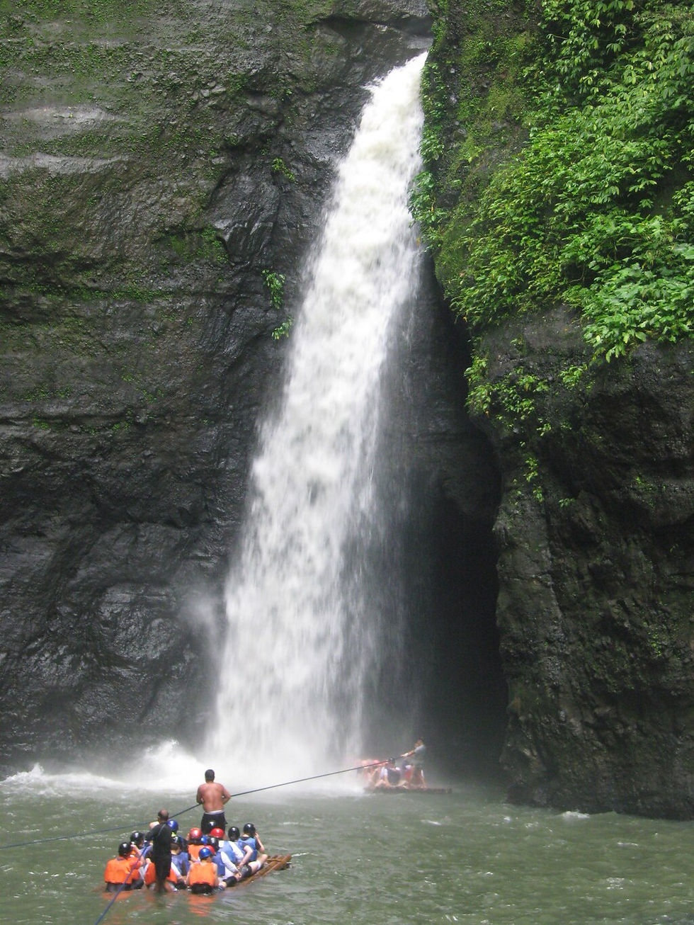 People in helmets and life vests raft near a tall waterfall amid lush green cliffs. The scene is dynamic and adventurous.