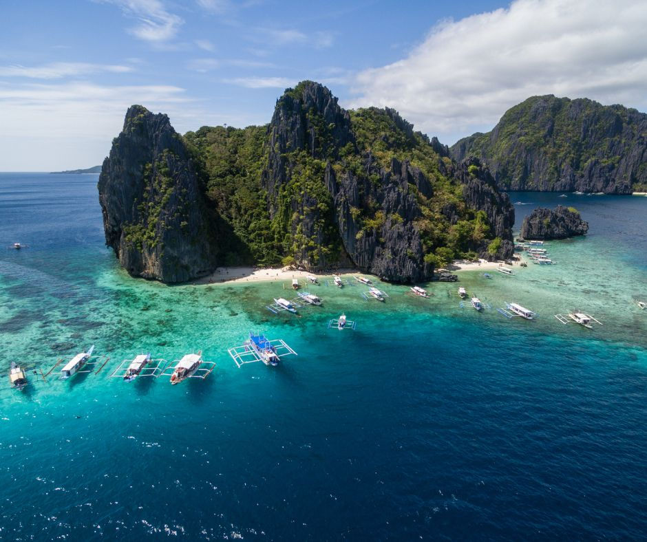 El Nido, boats float on turquoise waters by a lush, rocky island under a clear sky. White sand beach and calm, serene setting.